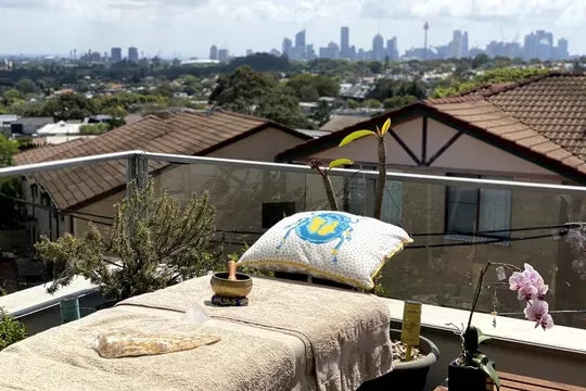 Massage table set on a balcony overlooking the Sydney CBD skyline, adorned with a beetle cushion, a majestic owl wing, a Tibetan bowl, and a blooming purple-pink orchid, ready for a transformative Reiki and Light Language experience, creating a serene and luxurious atmosphere